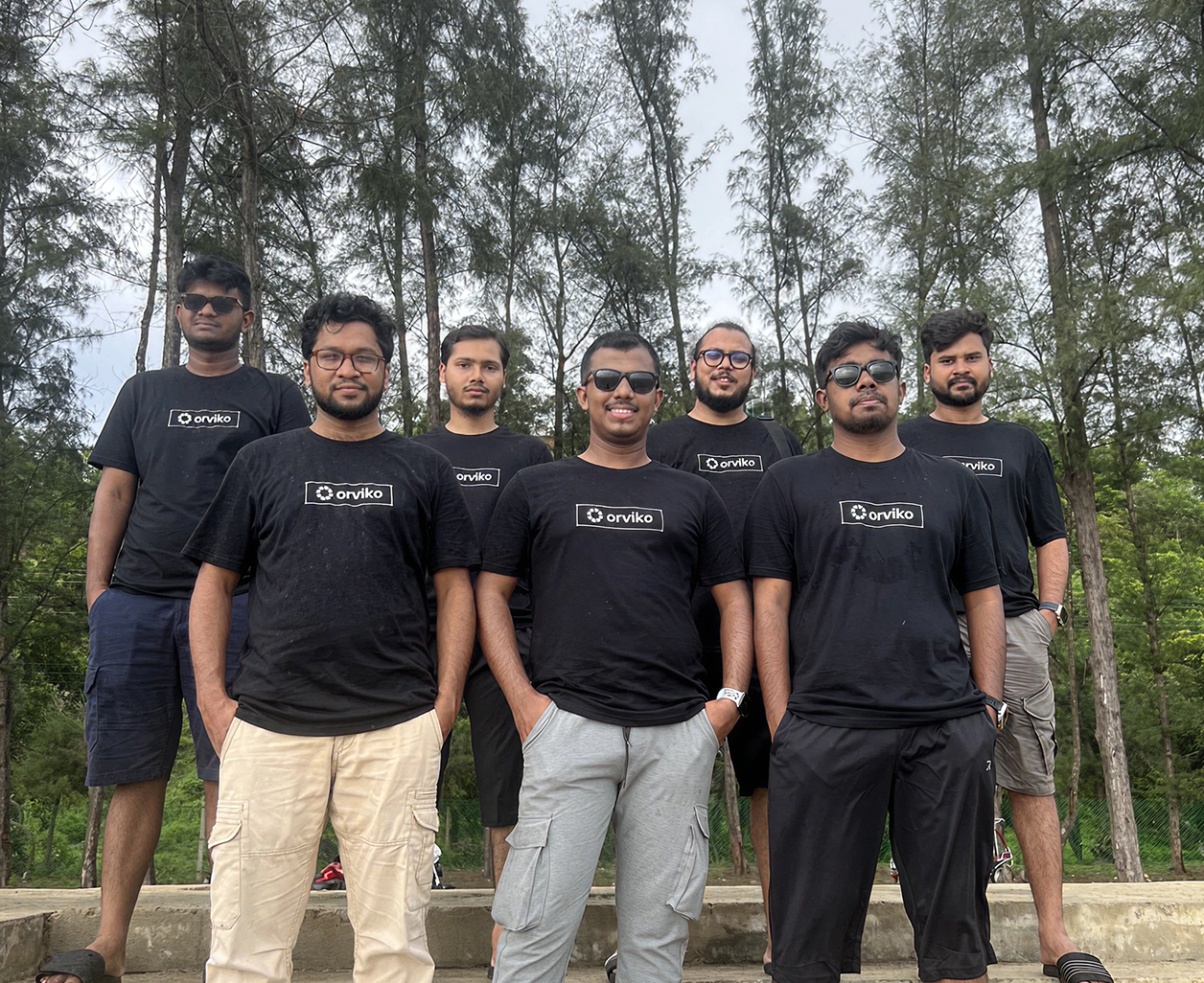 Group of seven men in black "orviko" t-shirts posing outdoors in front of trees.